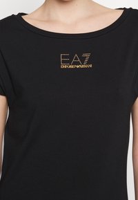 Czarna koszulka z szerokim dekoltem i krótkimi rękawami. Na przodzie znajduje się złote logo "EA7 EMPORIO ARMANI" ozdobione kryształkami.