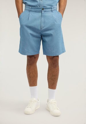 Uomo che indossa pantaloncini azzurri chiari, calzini bianchi alti e scarpe da ginnastica bianche, in piedi con le mani in tasca.