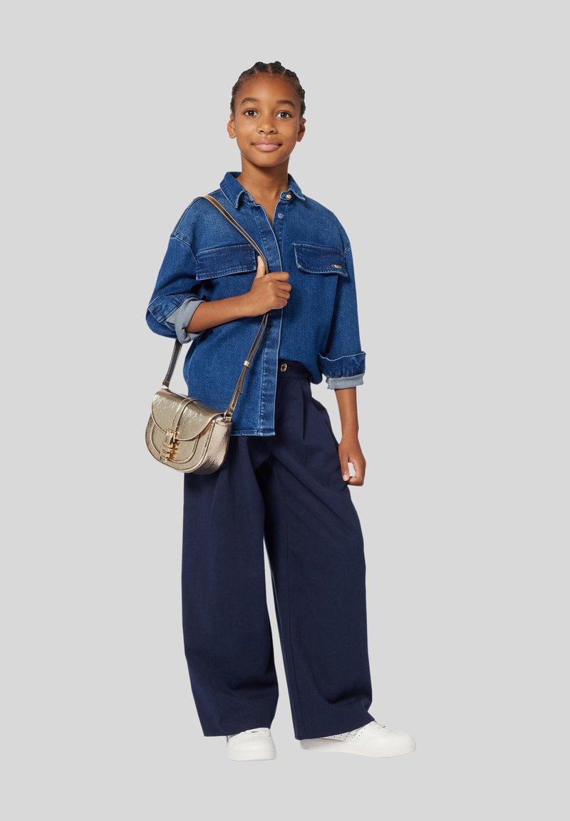 Denim oversized shirt, marineblauwe wijde broek, witte sneakers en een gouden crossbodytas. Huidtextuur is glad, haar is gestyled in vlechten.