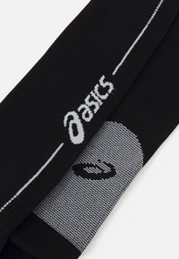 Sorte ankelstrømper med ribbet tekstur, der har et hvidt Asics-logo og en mønstret sektion i en lysere grå nuance.
