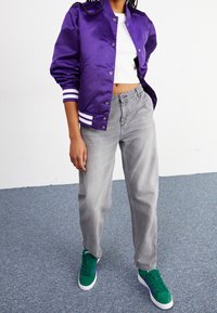 Paarse satijnen bomberjack met witte strepen, grijze losse jeans en groene sneakers met paarse accenten. Casual sportieve outfit detail.