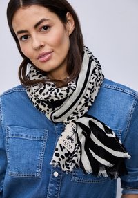 Femme aux cheveux foncés portant une écharpe à imprimé animal et rayée sur une chemise en denim bleu, sur un fond clair uni.