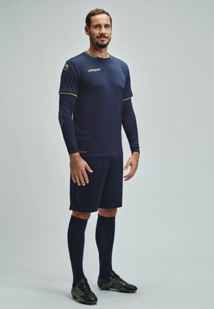 Uomo in piedi con uniforme da calcio blu scuro, con strato interno a maniche lunghe, calze alte scure fino al ginocchio e scarpe con tacchetti nere su sfondo grigio.