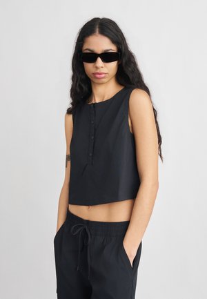 Ung kvinde med langt mørkt hår iført sort ærmeløs crop top, sorte bukser med snøre, sorte solbriller og næsering, stående med hænderne i lommerne.