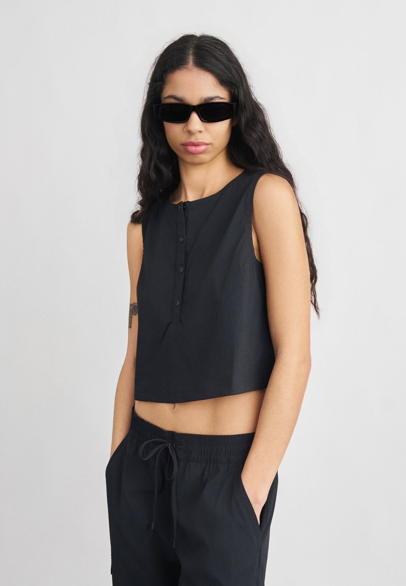 Νεαρή γυναίκα με μακριά σκούρα μαλλιά, ντυμένη με μαύρο αμάνικο crop top, μαύρο παντελόνι με κορδόνι, μαύρα γυαλιά ηλίου και δαχτυλίδι στη μύτη, στέκεται με τα χέρια στις τσέπες.