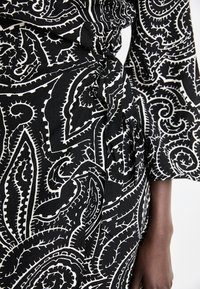 Abito in tessuto nero con motivo paisley bianco, caratterizzato da un dettaglio annodato in vita e maniche ampie. Materiale leggero e testurizzato.