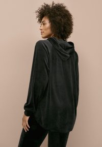 Zwarte velours hoodie met een zachte textuur, ontspannen pasvorm en een trekkoord kap. De hoodie heeft lange mouwen en een afgeronde zoom.