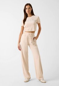 Beige cropped t-shirt with "GUESS" logo, paired with beige drawstring pants. Both pieces have a soft, smooth texture and relaxed fit.
