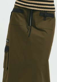 Falda midi verde oliva con un diseño estilizado. Presenta detalles de bolsillos negros y una cintura con cordón ajustable y cierre de cremallera.