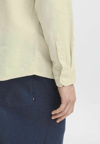 Helles beiges Langarmhemd mit geknöpften Manschetten, strukturiertem Stoff und abgerundetem Saum, kombiniert mit dunkelblauen Hosen mit Taschen-Details.