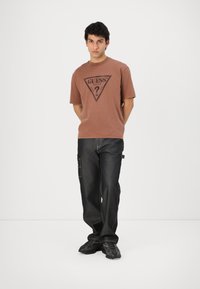 Brun kortermet t-skjorte med en svart trekantet logo, parret med mørke denim cargo-bukser med sidelommer og synlig søm.