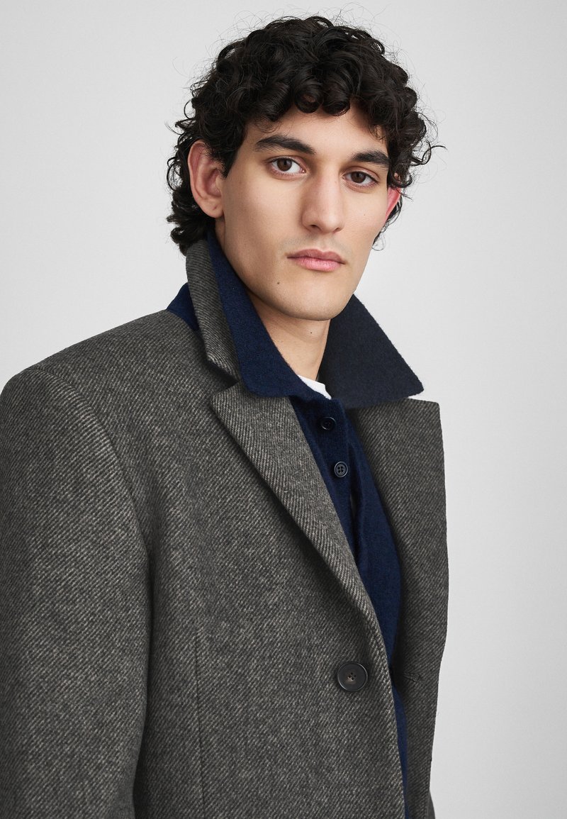 Homme aux cheveux bouclés et foncés portant un manteau en chevrons gris sur une chemise col bleu marine, regardant directement l'objectif devant un fond uni.