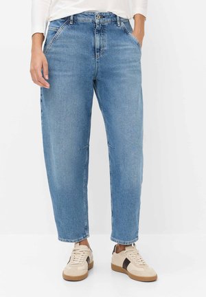 Person iført lyseblå jeans med afslappet pasform og cremefarvede sneakers med brune accenter står mod en hvid baggrund.
