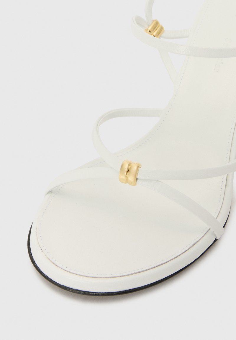 White strappy sandal featuring thin straps, gold hardware accents, a smooth leather texture, and a black sole edge.