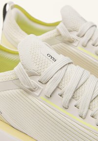 Fehér atlétikai cipők légáteresztő hálóból, texturált oldalpanelekkel, lapos fűzőkkel és a talp közelében finom sárga kiemeléssel.