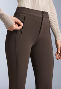 Hnědé slim fit kalhoty vyrobené z elastické látky, vybavené kapsami na zip po stranách a hladkým matným povrchem.