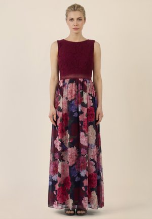 Frau in einem ärmellosen burgunderroten Kleid mit Spitzenoberteil und langem floralen Rock, stehend vor einem schlichten beigefarbenen Hintergrund.