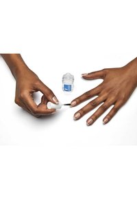 Application de vernis à ongles transparent avec un capuchon blanc, sur une main au teint moyen et des ongles naturels soigneusement finis. Petite bouteille étiquetée en bleu à proximité.