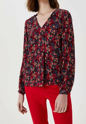 Vrouw die een lange mouwen bloemenblouse draagt met rode, beige en groene patronen, gecombineerd met felrode broek.