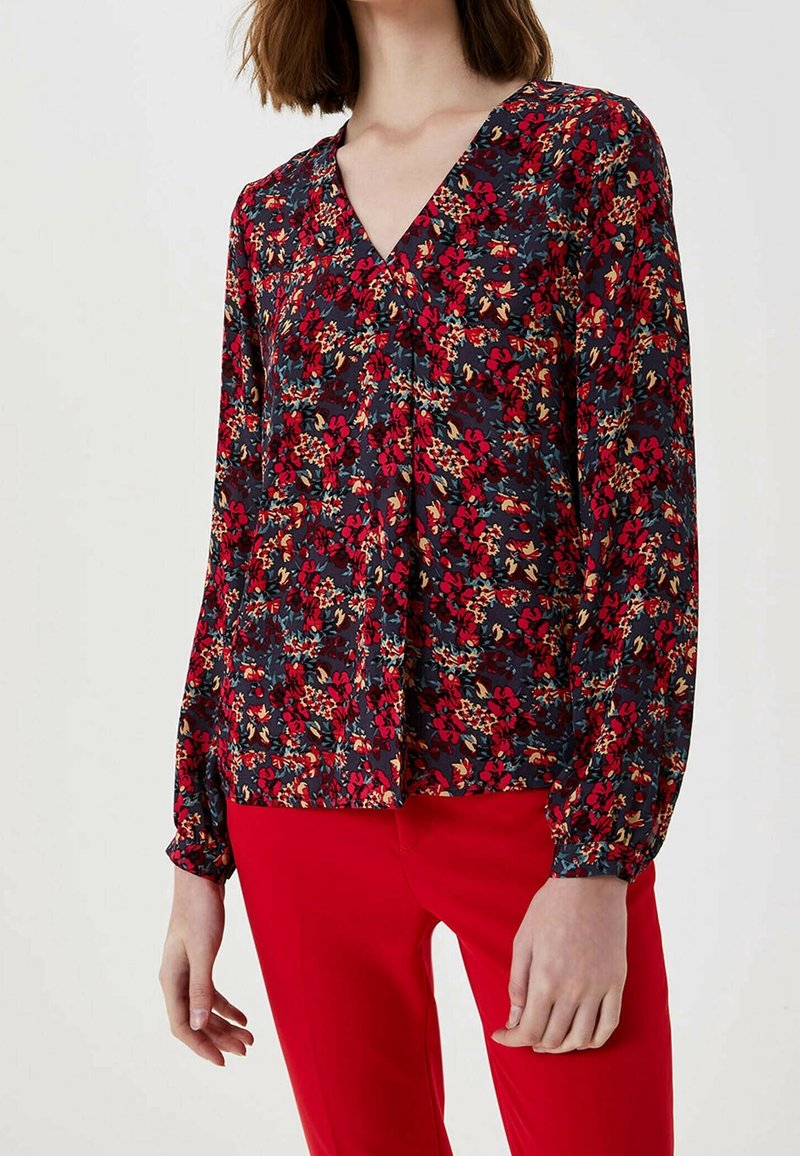 Femme portant un chemisier à manches longues à motifs floraux rouges, beiges et verts, associé à un pantalon rouge vif.