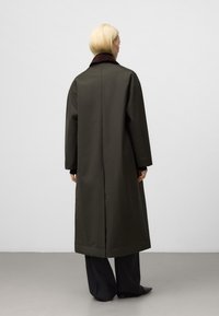 Cappotto lungo di colore verde oliva realizzato in tessuto liscio, con colletto scuro e design a taglio dritto. Silhouette minimalista senza decorazioni visibili.