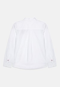 Bela srajca z dolgimi rokavi, ki vključuje ovratnik s gumbi, podrobnost z gubami na hrbtu in gumbnimi zapestji ter majhnim logotipom na rokavu.