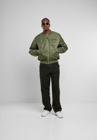 Giacca bomber verde oliva con zip, polsini e orlo a coste. Abbinata a pantaloni neri e sneakers bianche. Occhiali da sole completano il look.