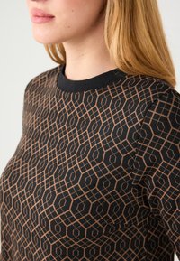 Haut noir à manches longues avec un motif géométrique hexagonal en marron clair. L'encolure ronde est bordée d'un bord noir uni. Tissu doux et texturé.