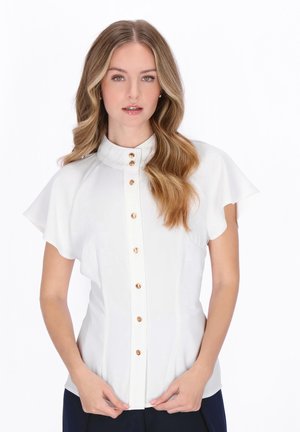 Młoda kobieta o falowanych blond włosach, ubrana w białą bluzkę zapinaną na guziki z krótkimi, falującymi rękawami, stojąca na tle jednolitego białego tła.