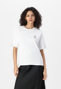 T-shirt blanc oversize à manches courtes, col rond, avec un logo brodé subtil en marron sur le côté gauche de la poitrine. Tissu en coton doux.