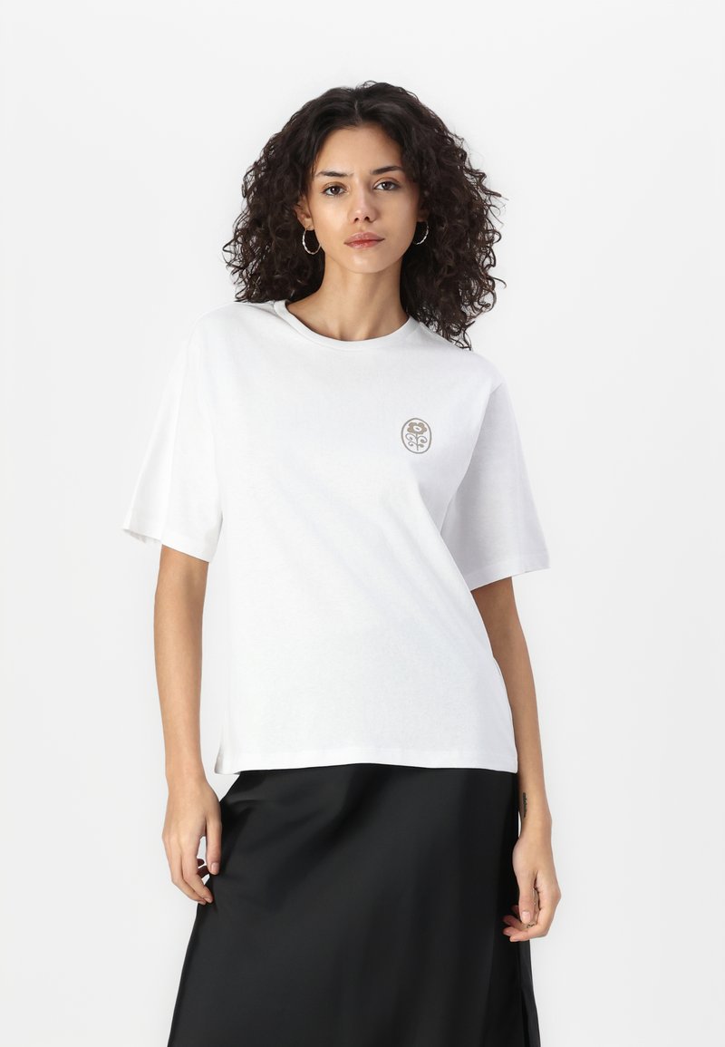 T-shirt blanc oversize à manches courtes, col rond, avec un logo brodé subtil en marron sur le côté gauche de la poitrine. Tissu en coton doux.