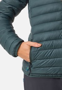 Gewatteerde jas van donkergroene stof, met horizontale stiksels, ritszakken en Zweedse vlagaccenten nabij de zoom.