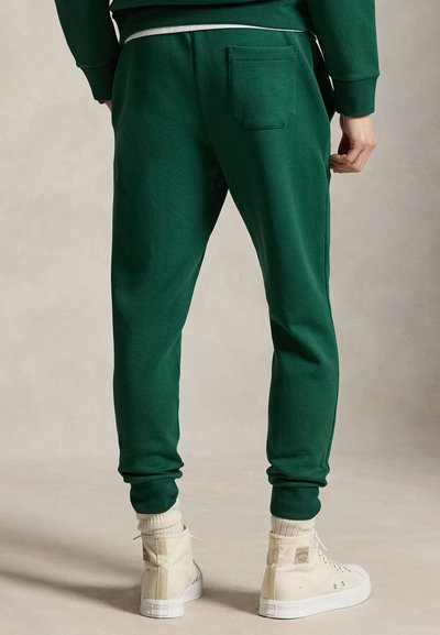 Persoon draagt donker groene joggerbroek met achterzak, crèmekleurige hoge sneakers en bijpassende sokken, staand op een neutrale achtergrond.