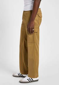 Pantaloni cargo khaki con gamba ampia, dotati di tasche laterali e di una texture liscia, abbinati a sneakers bianche e accenti scuri.