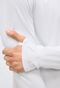 Venstre hånd som griper høyre overarm, iført hvit, teksturert langermet topp med tommelhull i mansjetten.