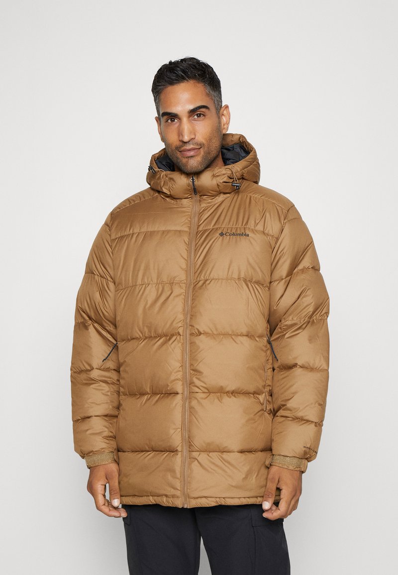 Mężczyzna w brązowej kurtce Columbia z ociepleniem i kapturem, stojący na tle jasnoszarym.
