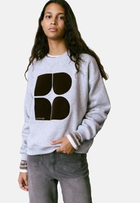 Sudadera gris con gráfico abstracto negro, puños acanalados con rayas marrones y blancas, combinada con pantalones grises.