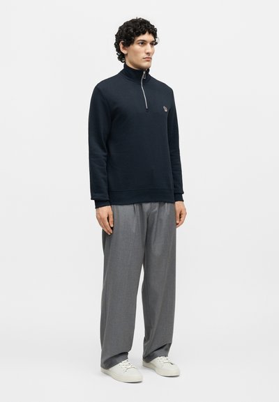 Άνδρας με σγουρά μαλλιά, φοράει μαύρο πουλόβερ με φερμουάρ στο λαιμό, γκρι φαρδιά παντελόνια και λευκά αθλητικά παπούτσια, στέκεται μπροστά σε απλό φόντο.