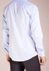 Světle modrá košile zobrazená zezadu, s dlouhými rukávy, hladkou texturou a zakřiveným lemováním. Nositelná s hnědými károvanými kalhotami.