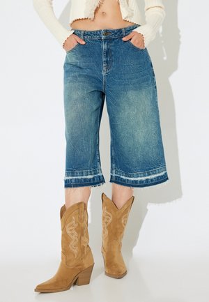 Culottes de denim con un degradado de lavado de claro a oscuro, dobladillo deshilachado y dos bolsillos laterales, combinados con botas de cowboy de ante en tono marrón.