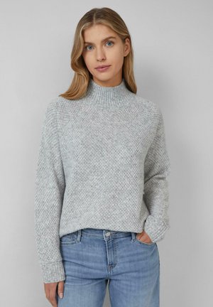 Jonge vrouw met lichtblond haar, gekleed in een lichtgrijze gestructureerde trui en blauwe spijkerbroek, staat tegen een effen grijze achtergrond.