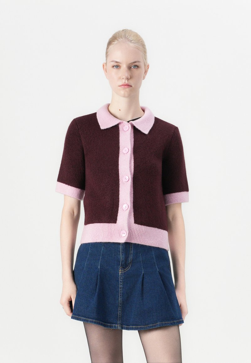 Short-sleeve cardigan in burgundy and pink with a soft collar, featuring six pink buttons and a textured finish. Paired with a denim mini skirt.