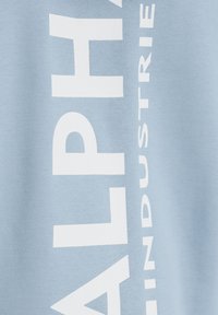 Svijetloplava dukserica od pamuka, s podebljanim bijelim natpisom "ALPHA INDUSTRIES" duž strane u vertikalnom rasporedu.