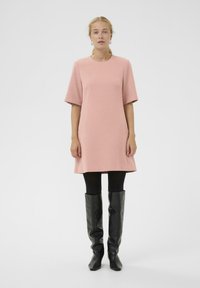 Rosa kurzärmeliges Kleid mit rundem Ausschnitt; kombiniert mit schwarzen Leggings und hohen, enganliegenden schwarzen Lederstiefeln. Glatte Textur, schlichtes Design.