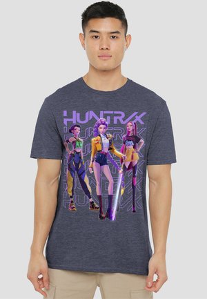 Young man wearing dark gray t-shirt featuring three animated female characters with futuristic outfits and neon purple "HUNTRX" text background.