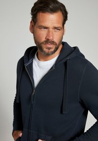 Marinefarbenes Zip-Hoodie aus weichem Stoff mit gefütterter Kapuze, seitlichen Taschen und weißen Stickdetails, getragen über einem weißen T-Shirt.