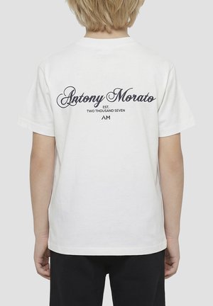 Bambino con capelli corti e biondi indossa una maglietta bianca con la scritta "Antony Morato EST. TWO THOUSAND SEVEN AM" sulla schiena, abbinata a pantaloni neri.