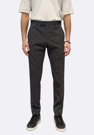 Uomo che indossa pantaloni aderenti grigio scuro, camicia bianca a maniche corte, sneakers nere con suola bianca e un braccialetto nero al polso sinistro.