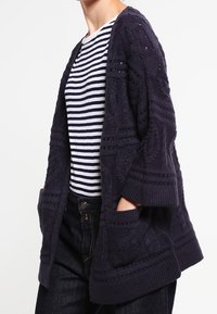 Mörkblått stickat kofta med kabelmönster, vida ärmar och två framfickor, lager över en randig skjorta.