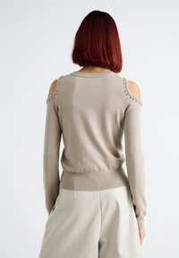 Maglione beige con design a spalle scoperte, orlo a coste e dettagli sulle spalle con borchie metalliche. Tessuto a maglia morbido.